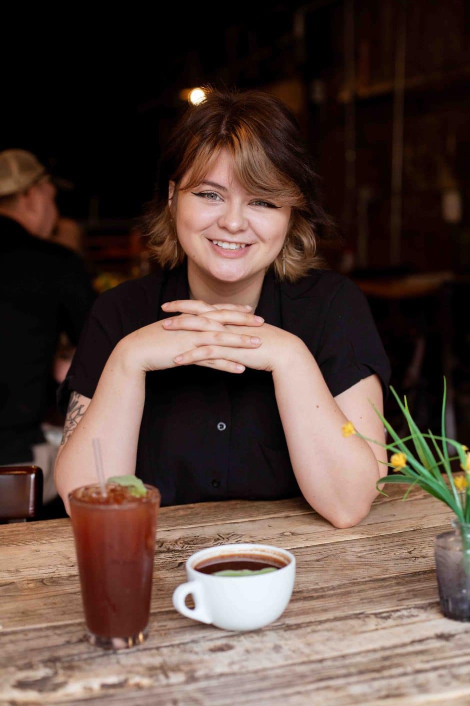 Person smiling and sitting at a wooden table with an iced drink and a hot drink in front of them. 