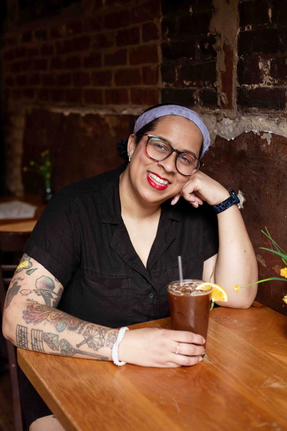 Barista Lilly Wilkenson sitting at a wooden table with an iced drink in their hands, smiling at the camera. 